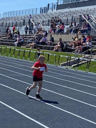 A young female student athlete wearing a red t-shirt and competition bib sprints down a blue all-weather track. Spectators watch from the bleachers in the background on a bright, sunny day.